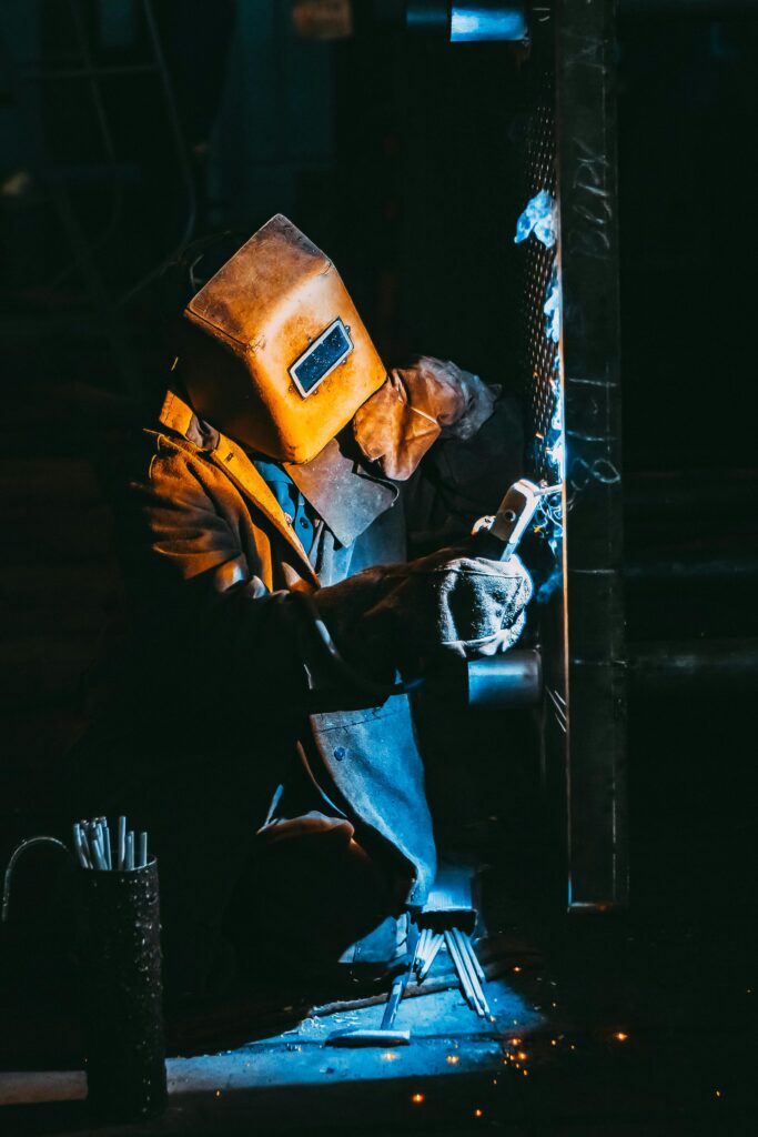 A welder wearing safety gear sparks metal in a dimly lit industrial setting.