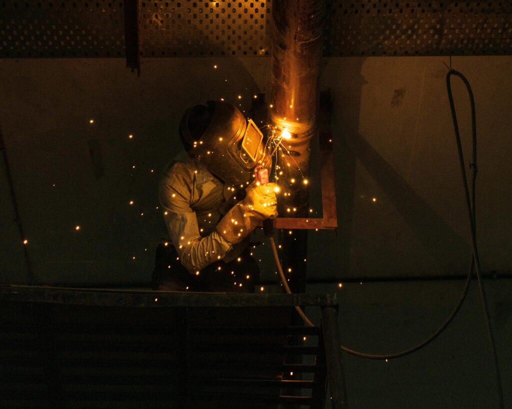 A welder at work indoors, creating bright sparks while welding metal pipes.